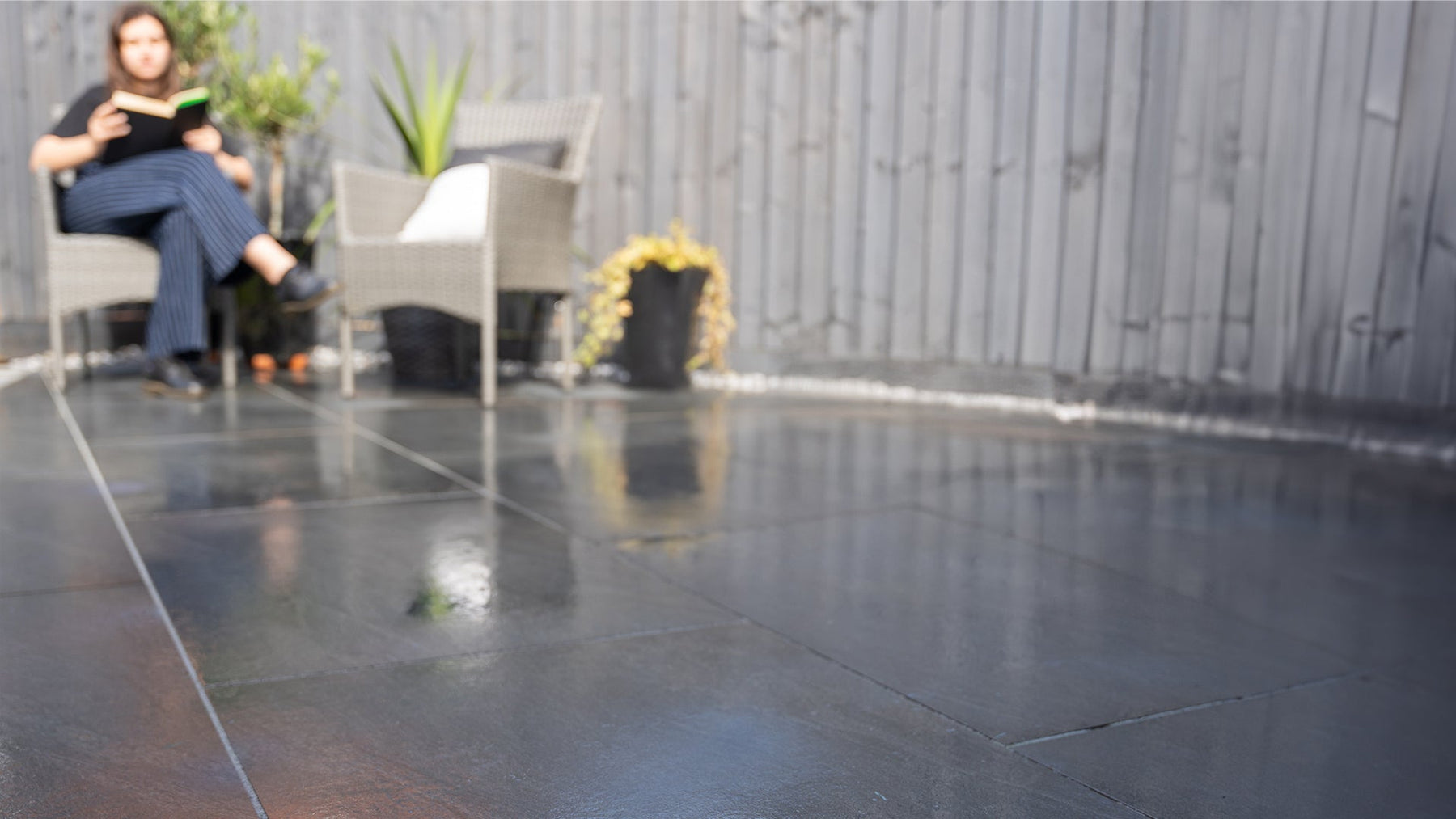 Garden photography, person reading book, sat in a garden where it has just rained, the Aravali Dark Grey porcelain paving are still wet from the rain. Installed by Up and Down Landscaping. Products provided by Select Living Group