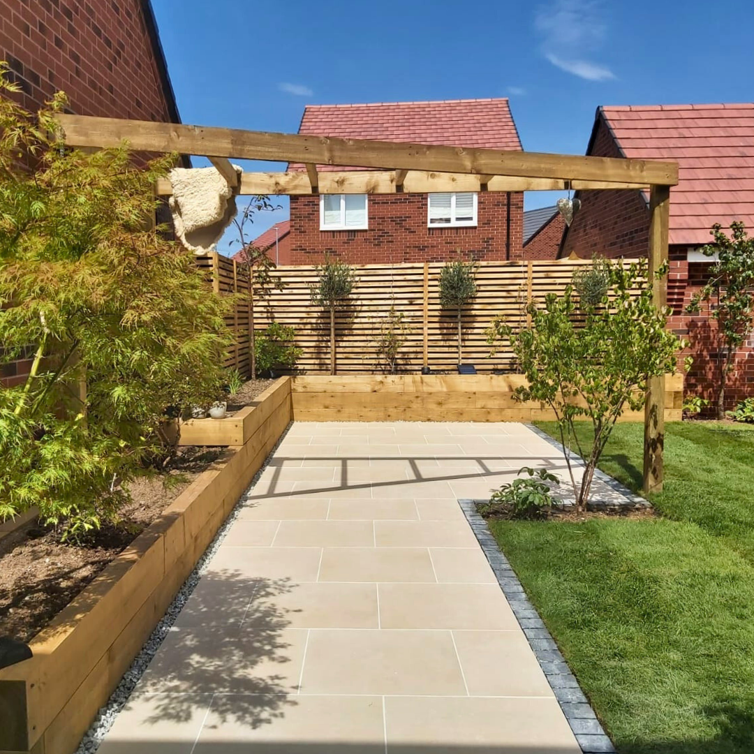 A Select Living Group beige porcelain paving stone installation in a garden setting with a wooden pergola, shrubbery, and a red-brick house in the background.