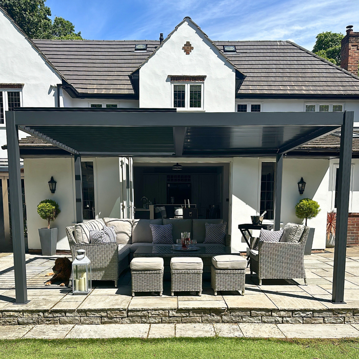 Select Living Group Aluminium garden pergola with a louved roof, installed over an outdoor seating area consisting of a sofa and chairs, set on a patio with a house in the background.