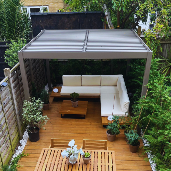 A 'Greige' aluminium garden pergola from Select Living Group with a louved roof, installed over a wooden deck area with outdoor furniture and potted plants.