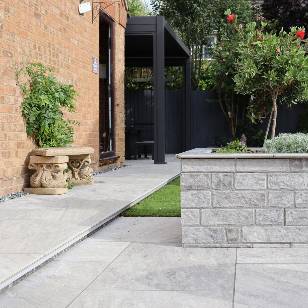 An outdoor setting featuring Select Living Group light grey porcelain paving with a garden and a brick wall in the background.
