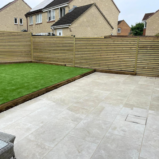 A backyard patio area featuring beige Porcelain Paving and Artificial Grass from Select Living Group with a residential fence and house in the background.