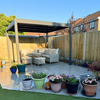 Aluminium garden pergola from Select Living Group with a two-part louvred roof, situated on a patio with outdoor furniture and potted plants in the background.